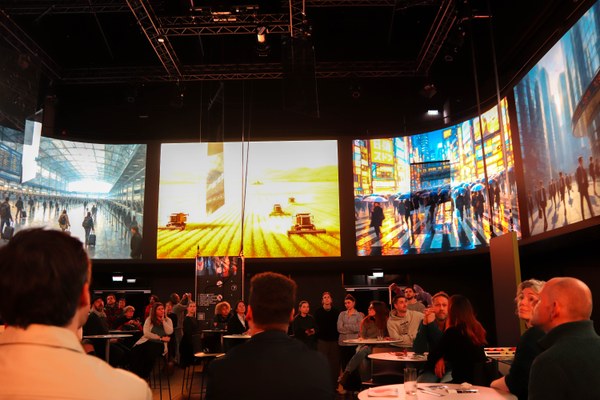 Besucherinnen betrachten in einem abgedunkelten Raum großflächige Leinwände mit farbenfrohen Stadt-, Bahnhof- und Agrarszenen während der Veranstaltung Lucid Dreams Echoes im Jahr 2025.