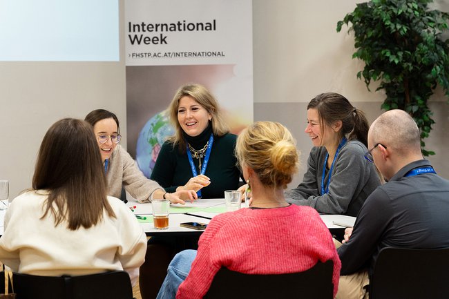 Gruppe von Personen sitzt an einem Tisch in einem Seminarraum und diskutiert während der International Week. Auf dem Tisch liegen Unterlagen, Stifte und Getränke, im Hintergrund ein Banner mit der Aufschrift ‚International Week‘. Gruppe von Personen sitzt an einem Tisch in einem Seminarraum und diskutiert während der International Week. Auf dem Tisch liegen Unterlagen, Stifte und Getränke, im Hintergrund ein Banner mit der Aufschrift ‚International Week‘.