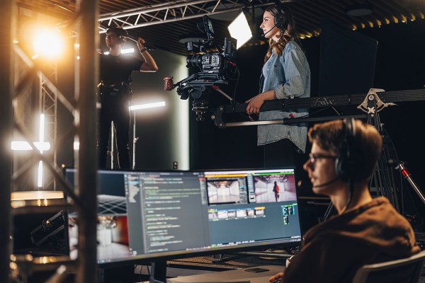 Arbeiten an einer Kamera auf einem Kranarm, während im Vordergrund Bildsignale und Video‑Software auf Monitoren bearbeitet werden.