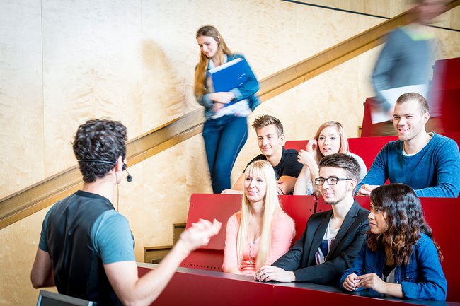 Im Audimax mit ansteigenden Sitzreihen: Vorne steht der Dozent mit Ansteckmikro, man sieht die ersten drei Sitzreihen, in denen Studierende sitzen und aufmerksam zuhören, zwei Personen sind auf dem Weg zu ihrem Platz