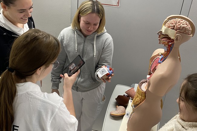 Studierende mit Smartphone bei scannen eines QR-Codes, welcher ihnen Aufgaben zum Lernen liefert. Davor ein Kunststofftorso eines Menschen.