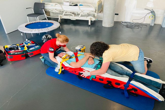 Auf dem Bild sieht man Stefan Rottensteiner in einer Rettungstrage am Boden liegen. Daneben sind zwei Studentinnen, die ihn verarzten.