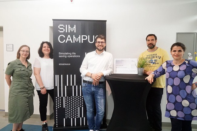 Auf dem Gruppenbild sieht man von links beginnend Lisa Brunhuber und Petra Ganaus stehend bei einem schwarzem SIM CAMPUS Banner. Gleich rechts daneben befindet sich Benjamin Roszipal, Manuel Kaider und Gabriella Szelesi neben einem Stehtisch, ebenfalls stehend.