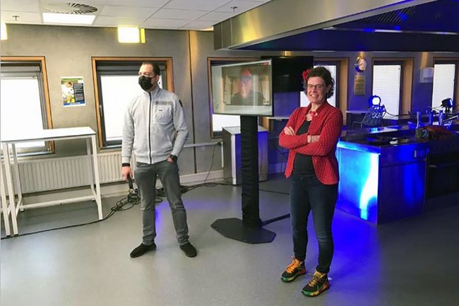 2 Vortragende, ein Mann und eine Frau, stehen seitlich vor einem Screen. Der Mann trägt eine FFP2 Maske und hält ein Mikrofon