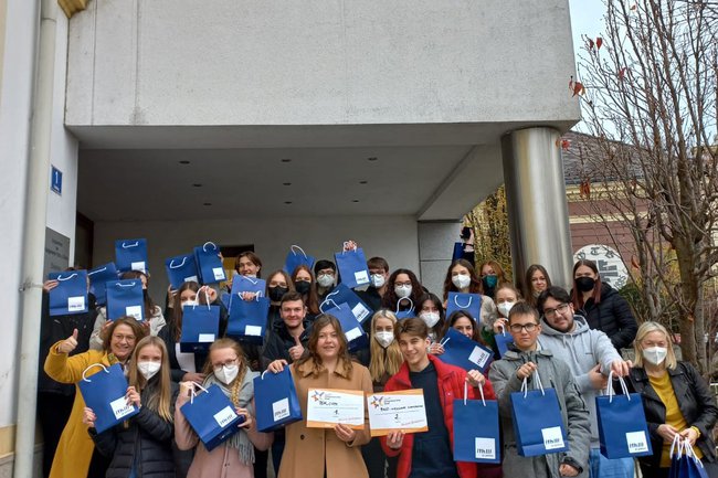 Wir sehen lachende Kinder, viele sind in den Masken. In den Händen tragen Kinder einen blauen FH Sackerl